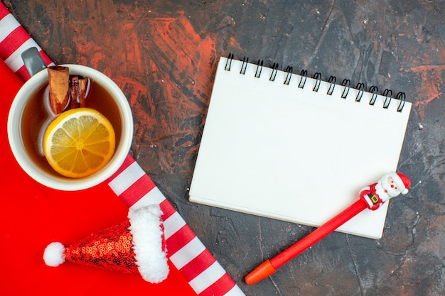 Vista dall'alto tazza di tè aromatizzata al limone e cannella mini taccuino cappello da Babbo Natale penna rossa su tovaglia rossa su tavolo rosso scuro