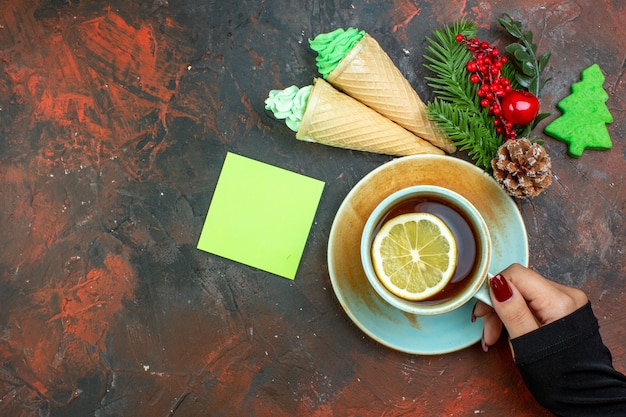 Vista dall'alto tazza di tè aromatizzata al limone in mano femminile ramo di albero di Natale gelati nota adesiva sul tavolo rosso scuro con posto libero