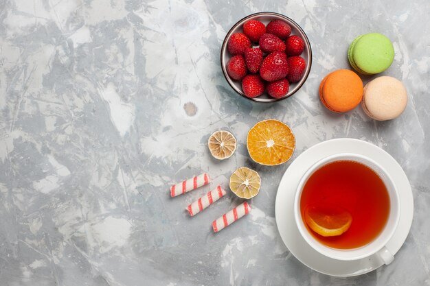 Vista dall'alto tazza di tè con fragole rosse fresche e macarons francesi su superficie bianca