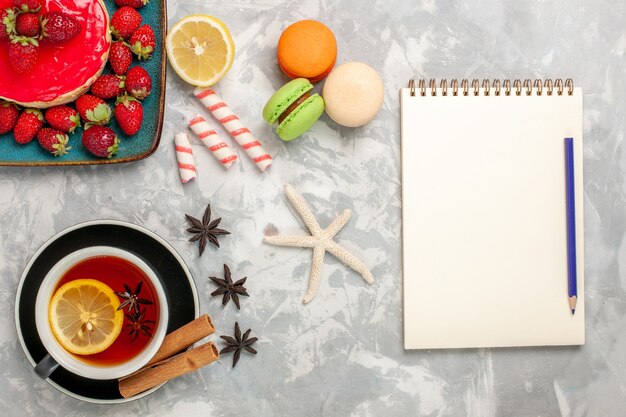 Vista dall'alto tazza di tè con macarons e piccola torta di fragole su superficie bianca