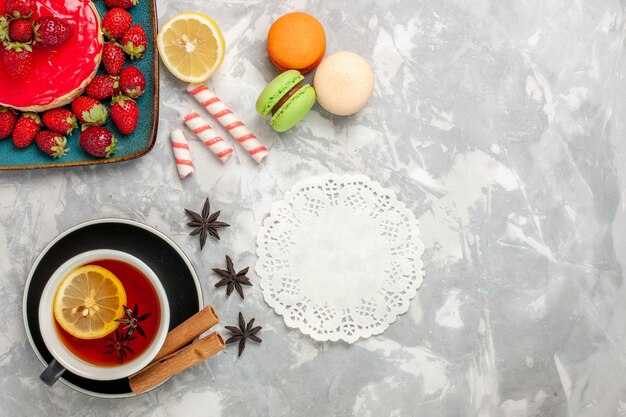 Vista dall'alto tazza di tè con macarons e piccola torta di fragole su superficie bianca