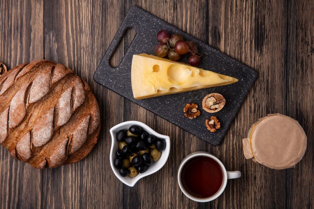 Vista dall'alto tazza di tè con olive in un piattino con formaggio maasdam su un supporto con yogurt e pane nero su uno sfondo di legno