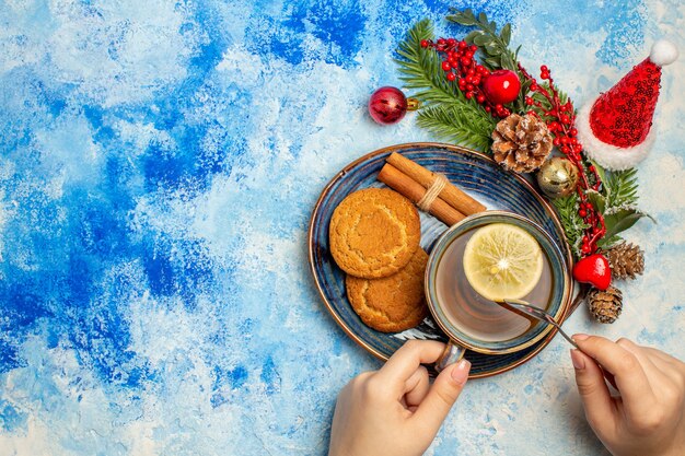 Vista dall'alto tazza di tè fette di limone bastoncini di cannella biscotti nel piattino ramo di albero di natale sul tavolo blu spazio libero