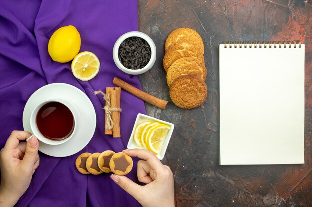 Vista dall'alto tazza di tè fette di limone in ciotola bastoncini di cannella tè in ciotola su tovaglia viola blocco note sul tavolo rosso scuro