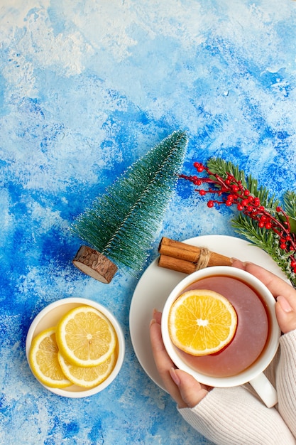 Vista dall'alto tazza di tè in mani femminili ramo di un albero di natale tagliare i limoni sul piattino piccolo albero di natale sul tavolo blu