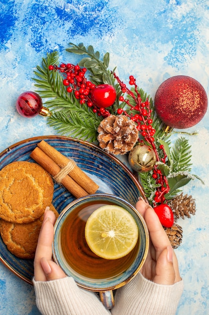 Vista dall'alto tazza di tè in mano donna fette di limone bastoncini di cannella biscotti in piattino sul tavolo blu