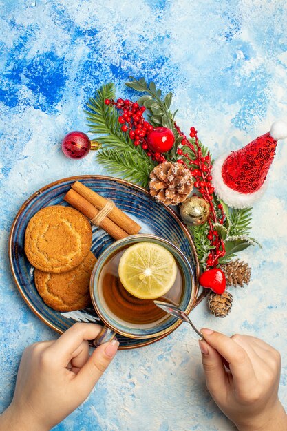 Vista dall'alto tazza di tè in mano femminile fette di limone bastoncini di cannella biscotti in piattino sul tavolo blu