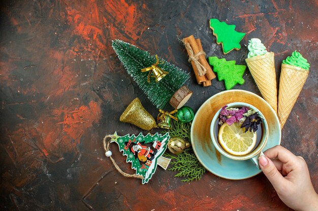 Vista dall'alto tazza di tè in mano femminile gelati bastoncini di cannella biscotti albero di natale ornamenti di natale sul tavolo rosso scuro