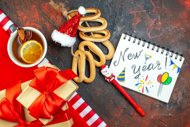 Vista dall'alto tazza di tè mini santa cappello su tovaglia rossa blocchi di legno bagel ovali capodanno scritto sui regali del blocco note sul tavolo rosso scuro