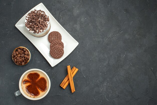 Vista dall'alto torta al cioccolato e biscotti su piatto rettangolare bianco tazza di caffè bastoncini di cannella ciotola con semi di caffè su sfondo scuro isolato