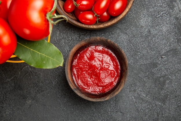 Vista dall'alto un secchio con pomodori e foglie di alloro ciotole con pomodorini e ketchup su fondo scuro