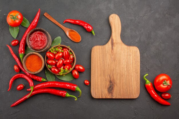 Vista dall'alto una ciotola di pomodorini peperoni rossi piccanti un cucchiaio di legno foglie di alloro ciotole di ketchup e polvere di peperoncino un tagliere e pomodoro su fondo nero