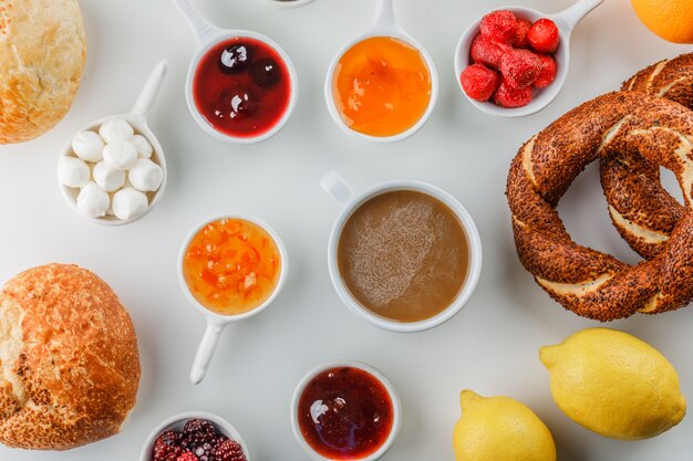 Vista dall'alto una tazza di caffè con marmellate, lamponi, zucchero, cioccolato in tazze, bagel turco, pane, limone su superficie bianca