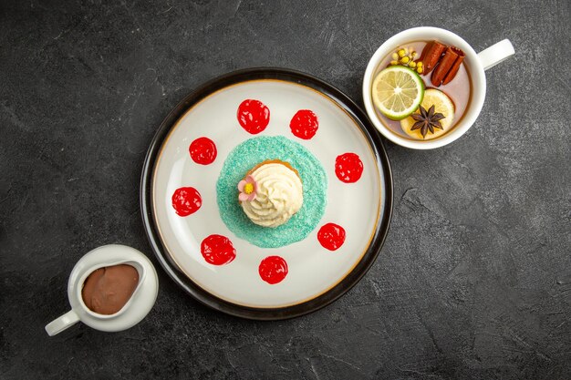 Vista dall'alto una tazza di piatto da tè di un appetitoso cupcake con salsa rossa una tazza di tè e una ciotola di crema al cioccolato sul tavolo nero