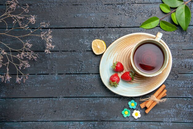 Vista dall'alto una tazza di tè e fragole sul piattino fetta di limone cannella caramelle fiore foglie sul tavolo di legno scuro