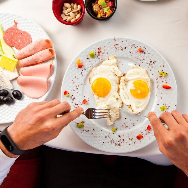 Vista dall'alto uova fritte sul tavolo una tovaglia bianca, un piatto con olive, formaggio, prosciutto, con noci, frutta candita mani di un uomo con una forchetta e coltello colazione