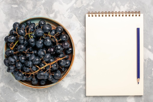 Vista dall'alto uva nera fresca succosa frutta dolce con blocco note sulla scrivania bianca chiara