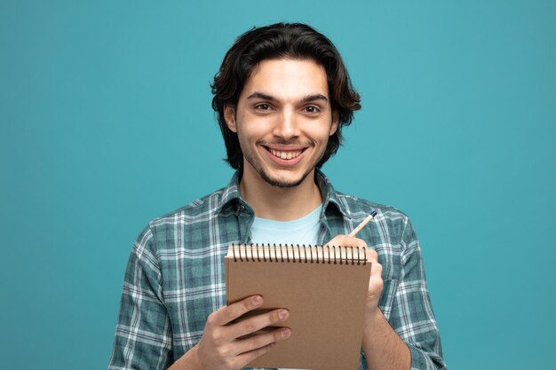 vista del primo piano del giovane uomo bello sorridente che tiene matita e blocco note guardando la fotocamera isolata su sfondo blu
