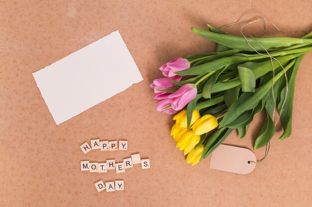 Vista dell&#39;angolo alto del testo felice della festa della mamma; carta bianca e gialla; fiori rosa tulipano sopra sfondo marrone