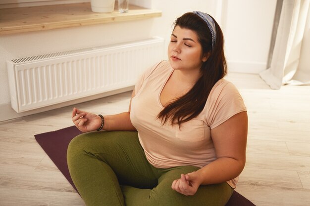 Vista di alto angolo di elegante giovane donna in sovrappeso paffuta vestita di leggings e t-shirt meditando con le gambe incrociate, chiudendo gli occhi, tenendosi per mano nel mudra, praticando tecniche di respirazione