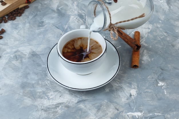 Vista di alto angolo versando il latte in una tazza di caffè con chicchi di caffè, bastoncini di cannella su sfondo grigio sgangherata. orizzontale