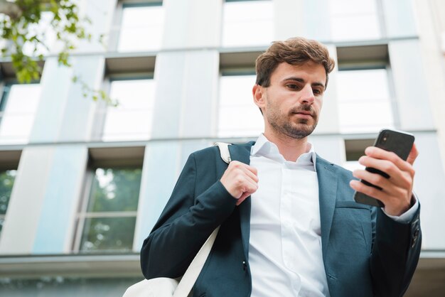 Vista di angolo basso di un uomo d&#39;affari che sta davanti a costruzione che esamina telefono cellulare