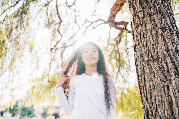 Vista di angolo basso di una ragazza sorridente che sta sotto l&#39;albero al sole