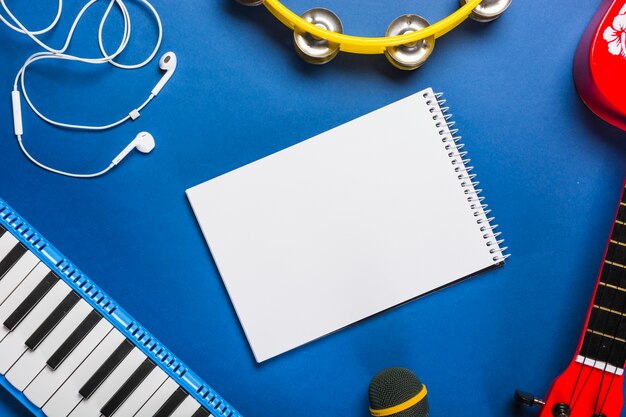 Vista elevata del blocchetto per appunti a spirale in bianco circondato con il trasduttore auricolare; chitarra; microfono; tastiera del pianoforte e tamburello su sfondo blu