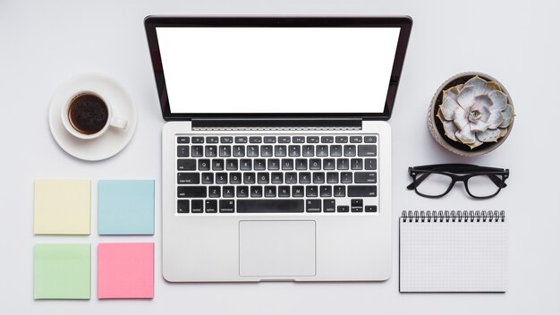 Vista elevata del laptop; note adesive; tazza di tè; blocco note a spirale; spettacoli e pianta succulenta su superficie bianca