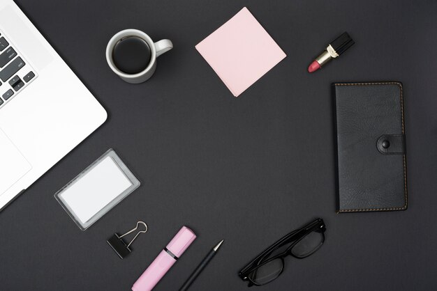 Vista elevata del laptop; rossetto; tazza di caffè e cancelleria per ufficio su sfondo nero