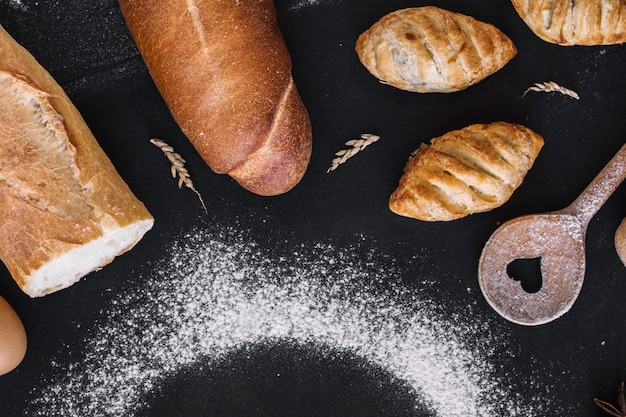 Vista elevata di pane fresco; cucchiaio a forma di cuore; grano e farina su sfondo nero