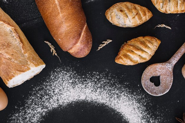 Vista elevata di pane fresco; cucchiaio a forma di cuore; grano e farina su sfondo nero