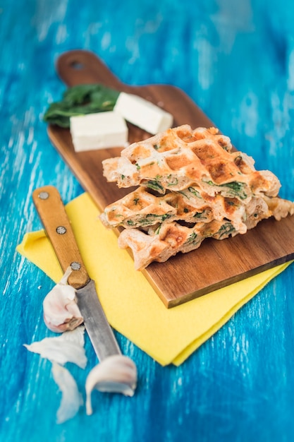 Vista elevata di waffles salati su tavola di legno con formaggio; chiodi di garofano di aglio e coltello su sfondo blu con texture