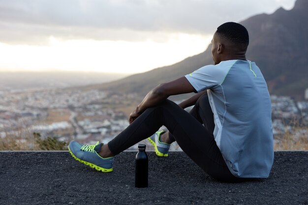 Vista esterna dell'uomo afroamericano rilassato con pelle scura e sana, beve acqua, si siede in collina, gode di un bellissimo paesaggio, atmosfera calma, montagne, vestito con abiti per lo sport. Benessere