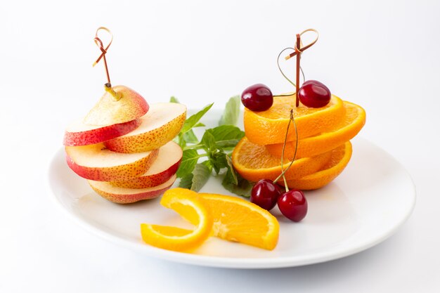 Vista frontale arance fresche affettate all'interno del piatto bianco con ciliegie rosse e pera su bianco