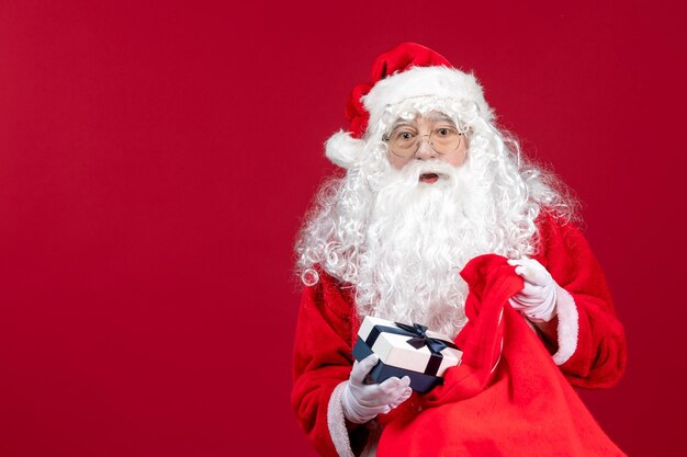 Vista frontale babbo natale che tiene presente dalla borsa piena di regali per i bambini sulla scrivania rossa capodanno emozione vacanza natale