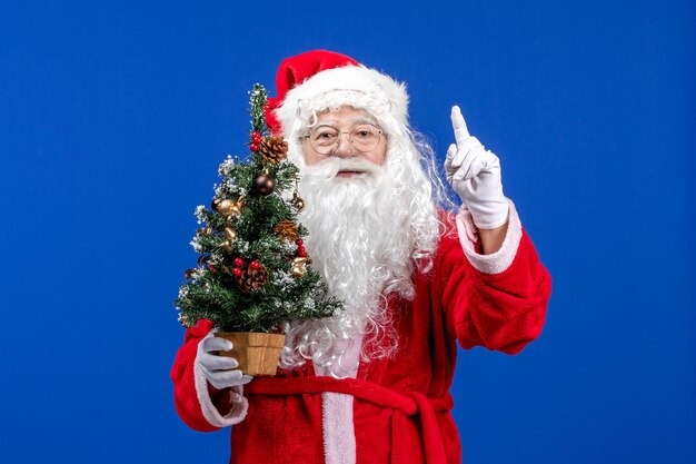 Vista frontale babbo natale che tiene un piccolo albero di capodanno sul pavimento blu neve colore natale capodanno