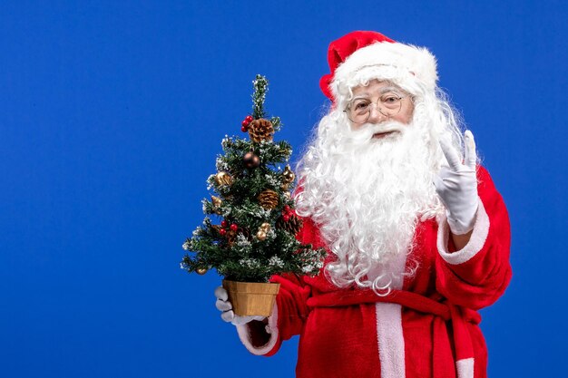 Vista frontale babbo natale che tiene un piccolo albero di capodanno sulla scrivania blu natale capodanno