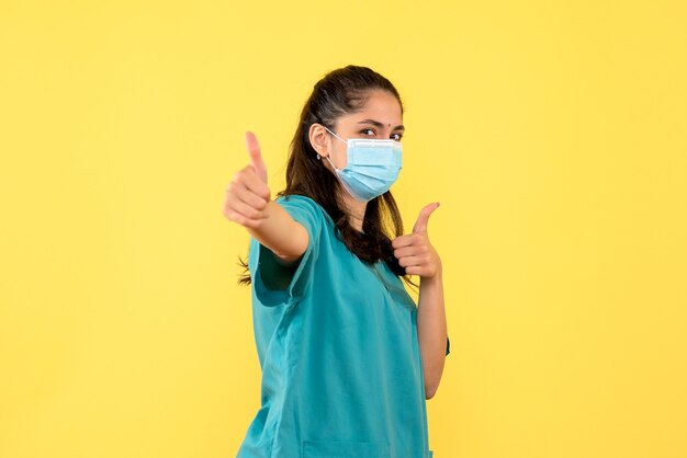 Vista frontale bella dottoressa in uniforme che fa pollice sul segno in piedi su sfondo giallo