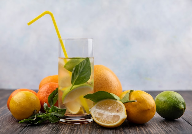 Vista frontale bicchiere d'acqua con fettine di limone lime e menta con paglia gialla e pompelmo con arance su fondo in legno