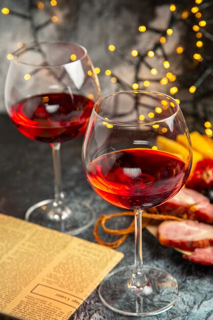 Vista frontale bicchiere di vino uva pezzi di formaggio fette di carne su piatto di legno giornale su luci di natale scure