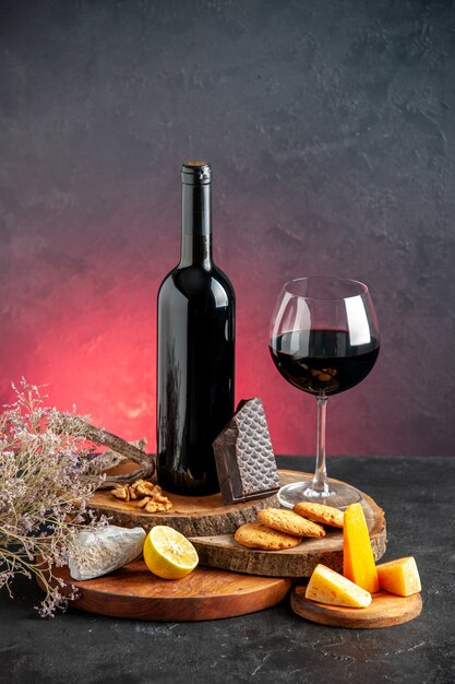 Vista frontale bottiglia di vino nero vino rosso in vetro formaggio tagliato limone pezzi di cioccolato fondente su tavole di legno ramo di fiori secchi sul tavolo rosso