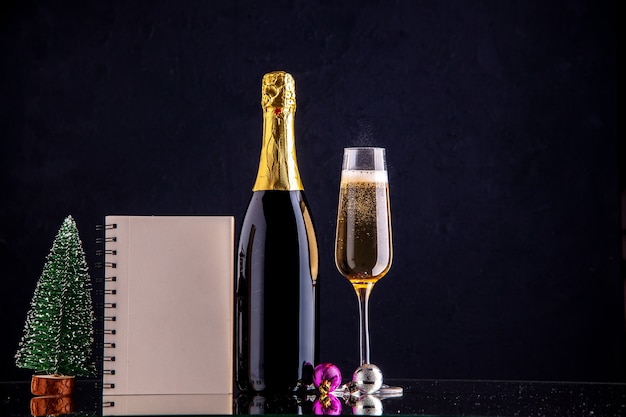 Vista frontale champagne in bottiglia e vetro mini taccuino albero di natale su superficie scura