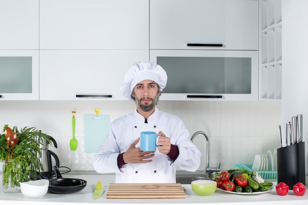 Vista frontale chef maschio serio in cappello da cuoco che tiene tazza in piedi dietro il tavolo della cucina