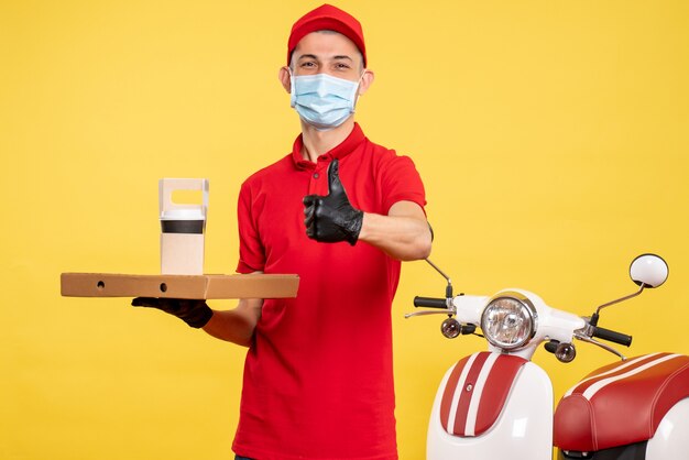 Vista frontale corriere maschio in maschera con consegna caffè e scatola su giallo lavoro colore servizio covid-virus pandemia uniforme da lavoro