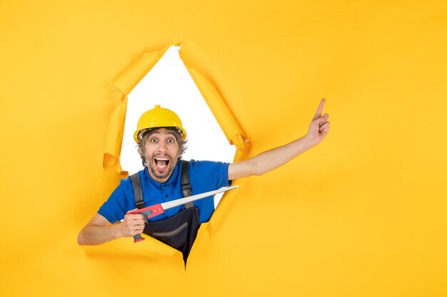 Vista frontale costruttore maschio in uniforme con strumento in mano sul muro giallo operaio edile lavoro costruttore lavoro colore