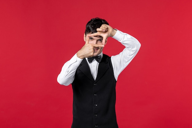 Vista frontale del cameriere maschio sorridente in uniforme con farfalla sul collo che fa un gesto ok su sfondo rosso