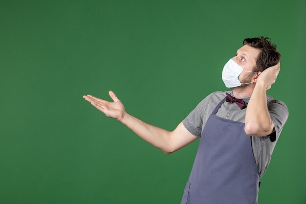 Vista frontale del cameriere scioccato in uniforme con maschera medica e rivolto verso l'alto sul lato destro su sfondo verde