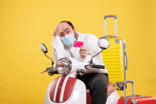 Vista frontale del concetto di viaggio con giovane ragazzo esausto in maschera medica seduto su una moto con valigia gialla su di esso e in possesso di carta di credito