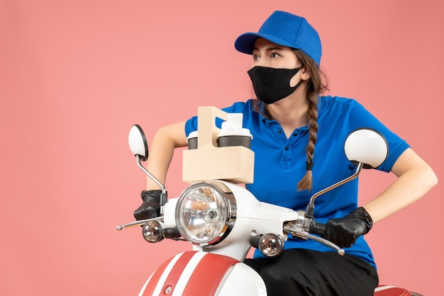 Vista frontale del corriere femminile concentrato che indossa maschera medica nera e guanti che tengono una piccola scatola su sfondo pesca